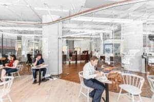 A modern office interior with people working at individual white tables, glass partitions, exposed columns, and ambient lighting.
