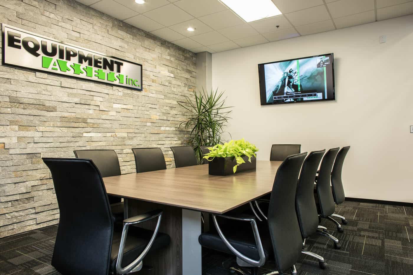Modern office meeting room featuring a long wooden table, black chairs, a wall-mounted tv displaying graphics, and the company's name "equipment assets inc." on a stone wall with green plants.