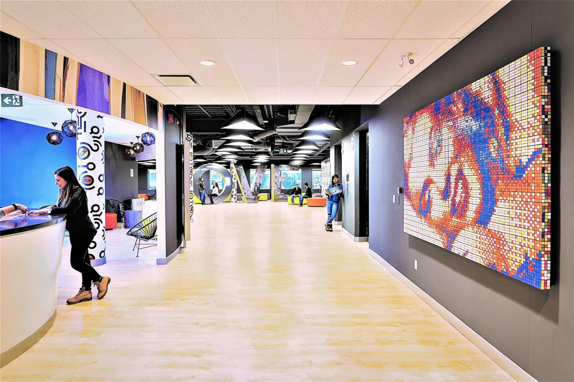 A modern office design and build with a vibrant, pixelated wall art, a front desk on the left with a receptionist, and a person walking down the corridor.
