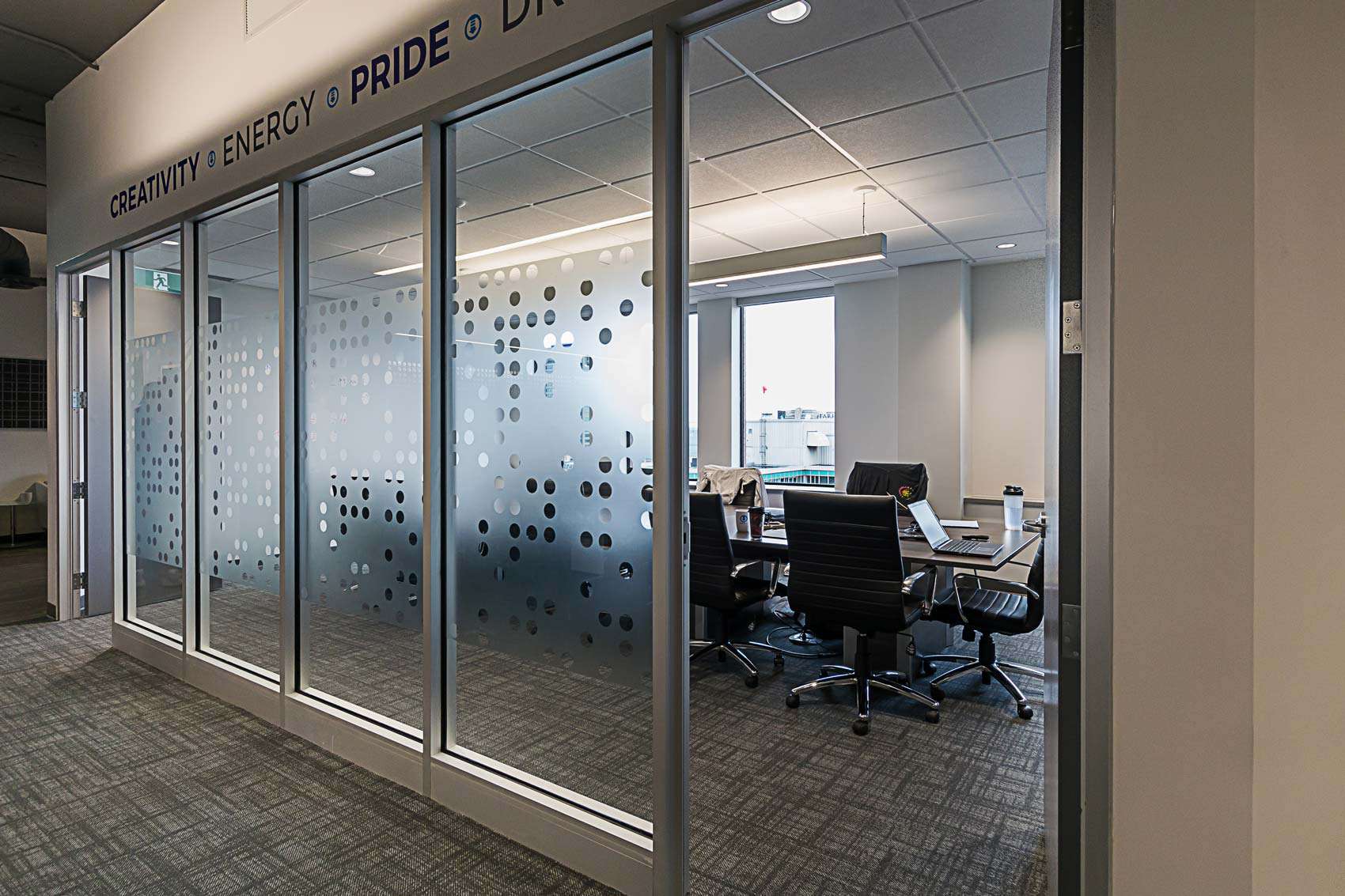 Modern office space visible through glass doors with the words "creativity, energy, pride, drive" written on top; features desks, chairs, and computers inside a brightly lit room.
