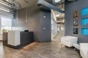 Modern office reception area with gray walls, a white desk, white sofas, pendant lights, and framed pictures on the wall.