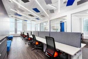 Modern office space with rows of desks and chairs, large windows, overhead lighting, and blue accents on walls and ceiling. Projects are displayed prominently throughout the area.