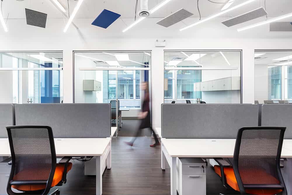 Modern office space featuring rows of desks with gray partitions, orange chairs, and projects displayed on monitors with a blurred figure walking past glass-walled rooms.