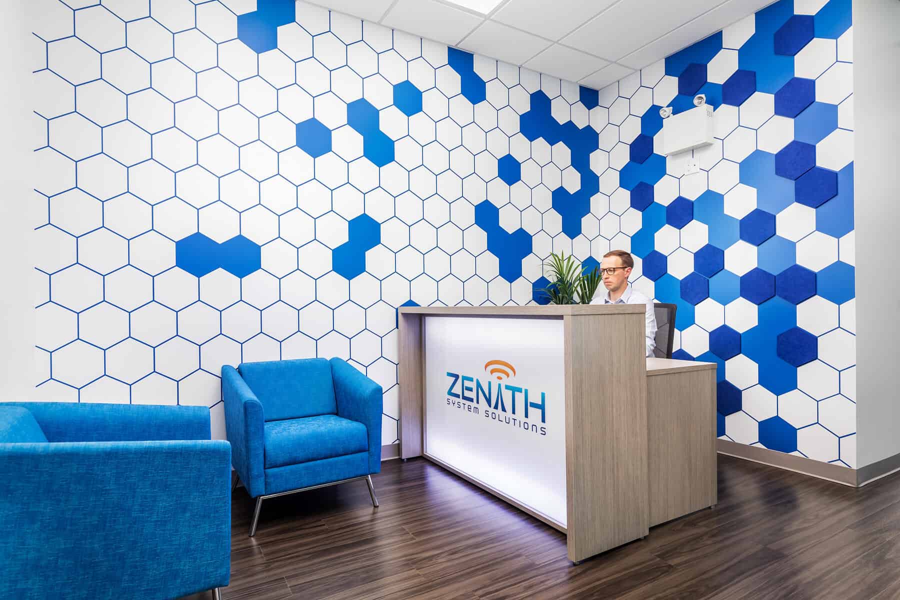 A reception area featuring a man behind a desk labeled "zenith laser solutions," with blue and white hexagonal patterned walls and two blue chairs.