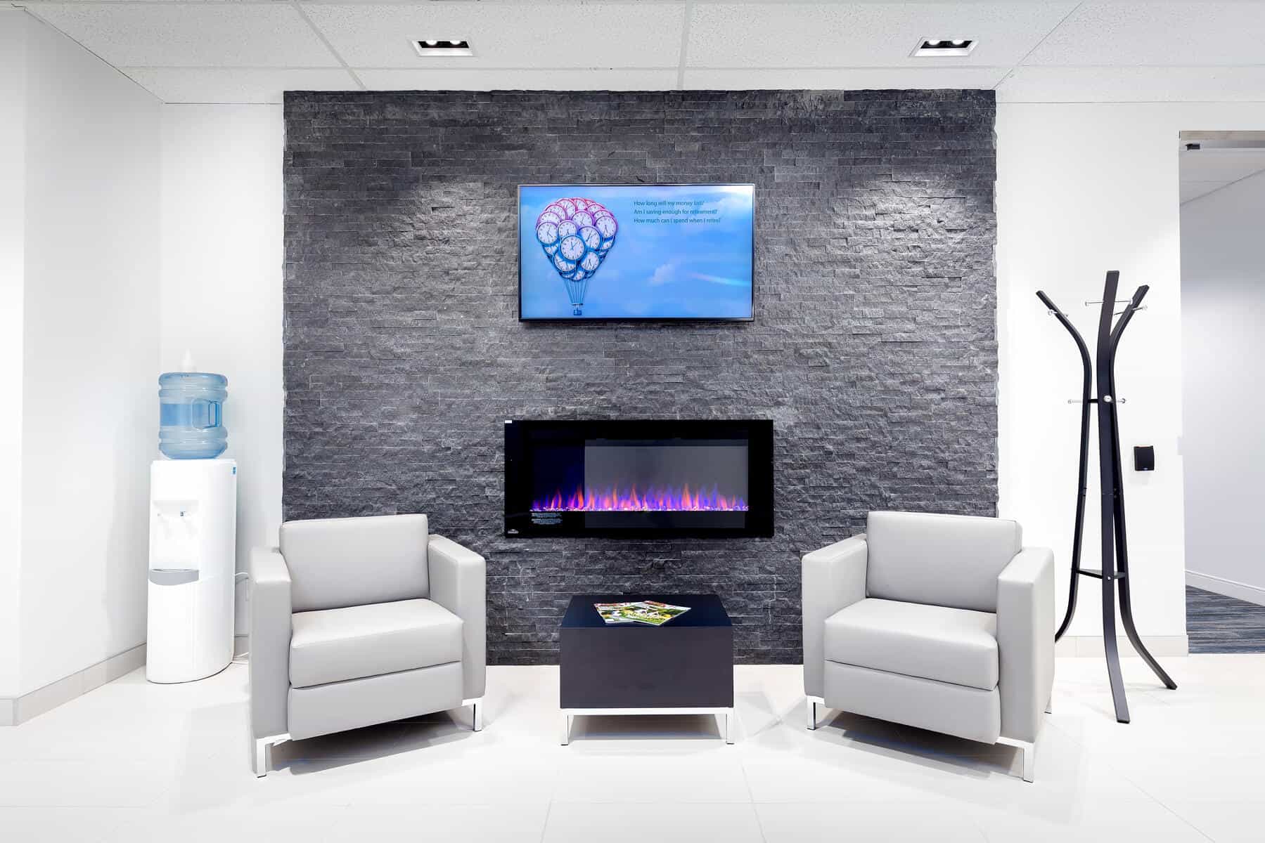 Modern office waiting area with two white chairs, a digital fireplace, a water cooler, coat stand, and an informative screen on a dark textured wall.
