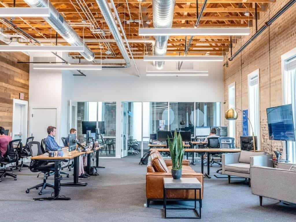 Modern office space designed with good office design principles, featuring employees working at desks with computers; includes exposed wooden beams, sofas, and a casual seating area.