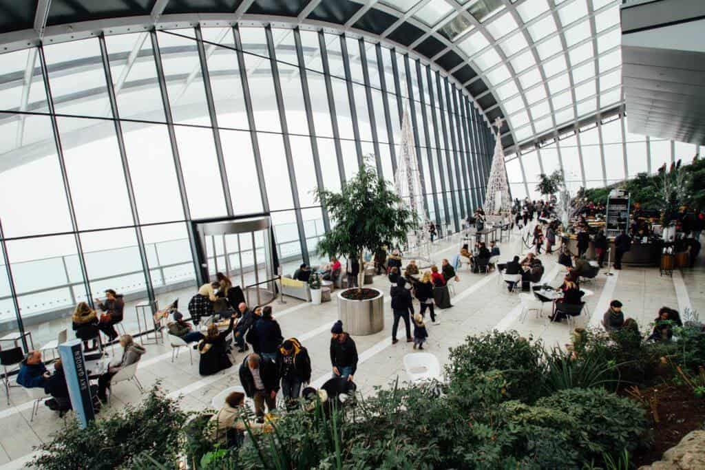 Interior of a modern airport terminal featuring a curved glass facade, with travelers seated and walking around indoor plants and cafes, reflecting emerging themes in post-pandemic workplace design.