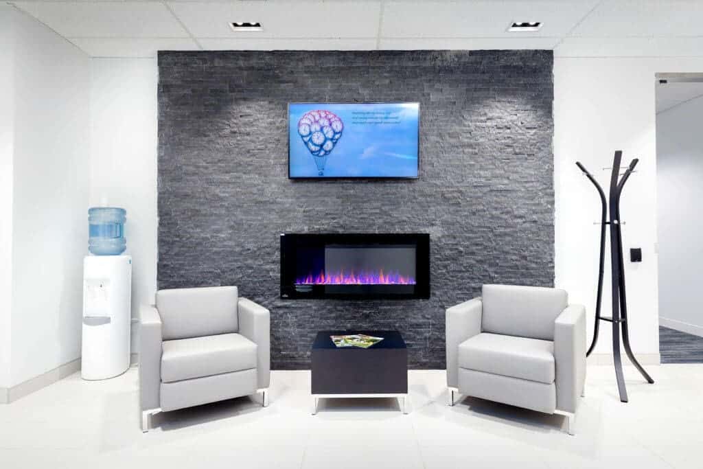 Modern office lobby designed for a successful hybrid workplace, featuring two white armchairs, a wall-mounted digital fireplace, a water cooler, and an informational screen on a textured grey wall.