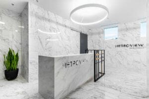 Modern, marble-clad reception area with a white desk labeled "metroview," circular overhead lighting, and potted plants, framed by two windows and minimalist decor.