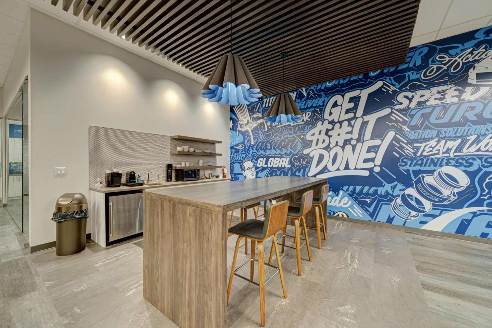 Modern office renovation kitchen and lounge area with wooden bar stools, a large blue and white mural with motivational text, and contemporary lighting.
