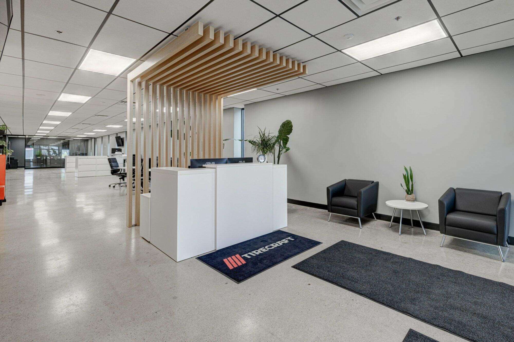 Modern office lobby with white reception desk, gray chairs, wooden slat design elements, projects showcased on the walls, and potted plants.