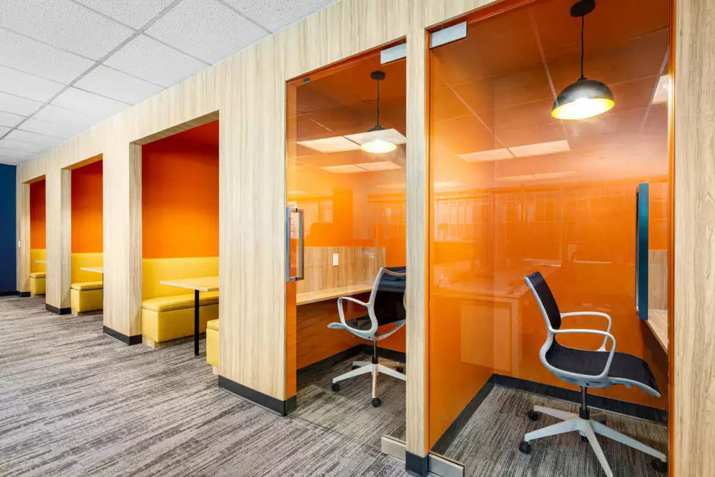 A row of booths with glass doors set into a wall, one with benches for meeting and two with task chairs and desks