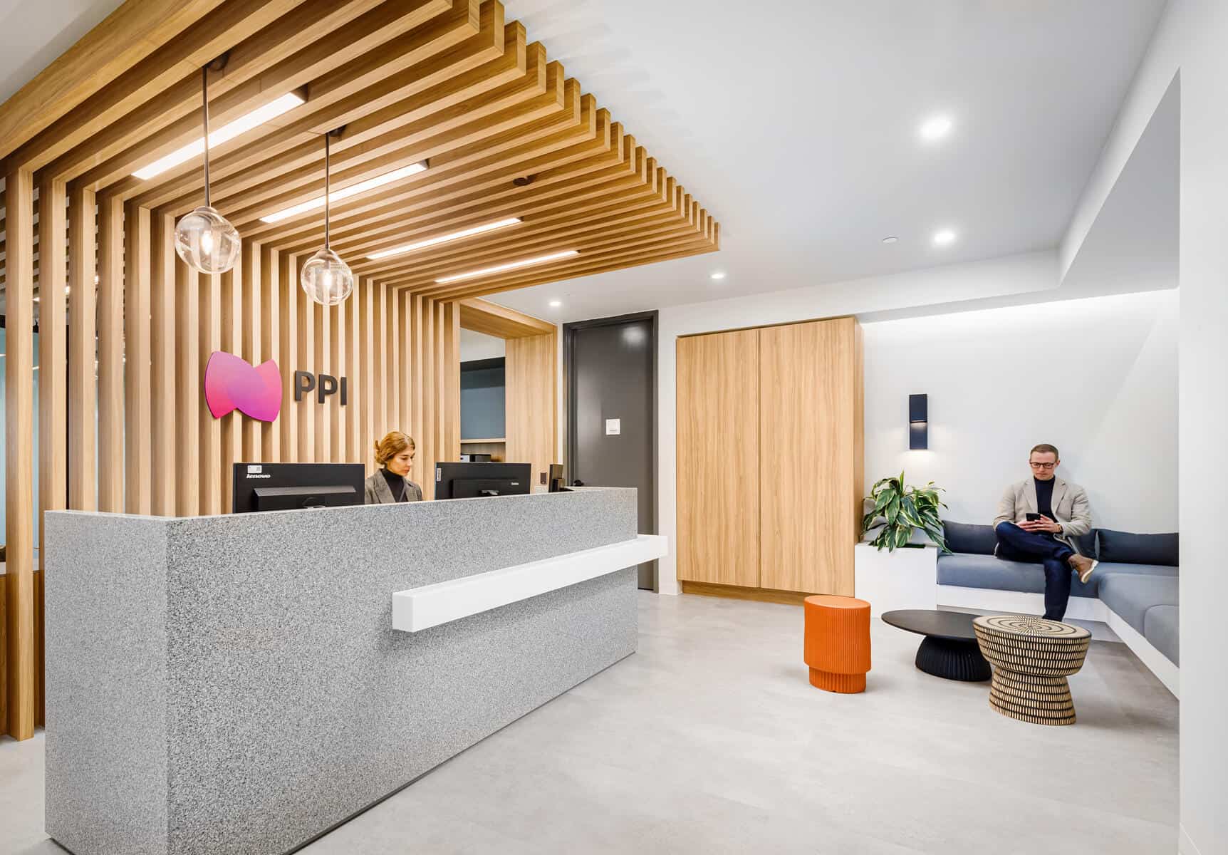 Modern office reception area with a man and a woman, stylish wooden accents, entrance desk, seating area, and hanging lights following an office renovation.