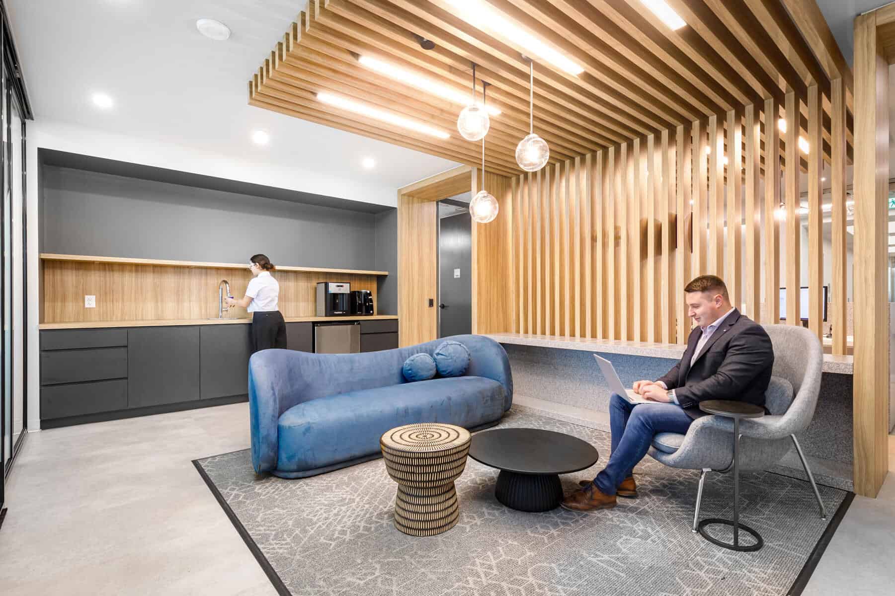 Modern office reception area with wooden slats on the ceiling, a blue sofa, and a man sitting in a chair working on a laptop. A woman stands at the counter in the background. The space