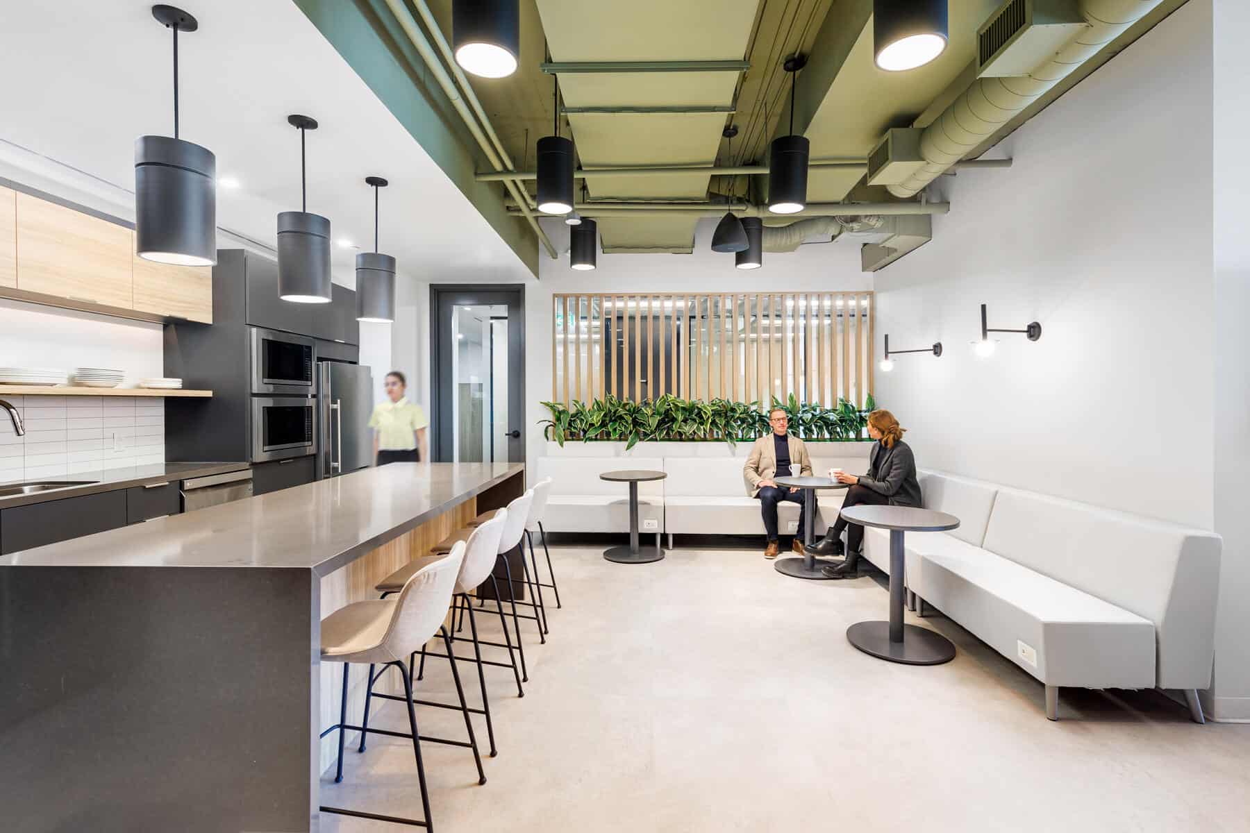 Modern office kitchen and break area, recently renovated with employees enjoying the new setup that includes a high bar table, stools, kitchen counter, and a relaxed seating area.