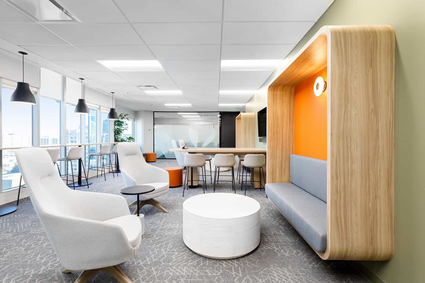 Modern office lounge area designed for an office renovation, with wooden booth seating, white chairs, a round table, and a background of high stools by the window.