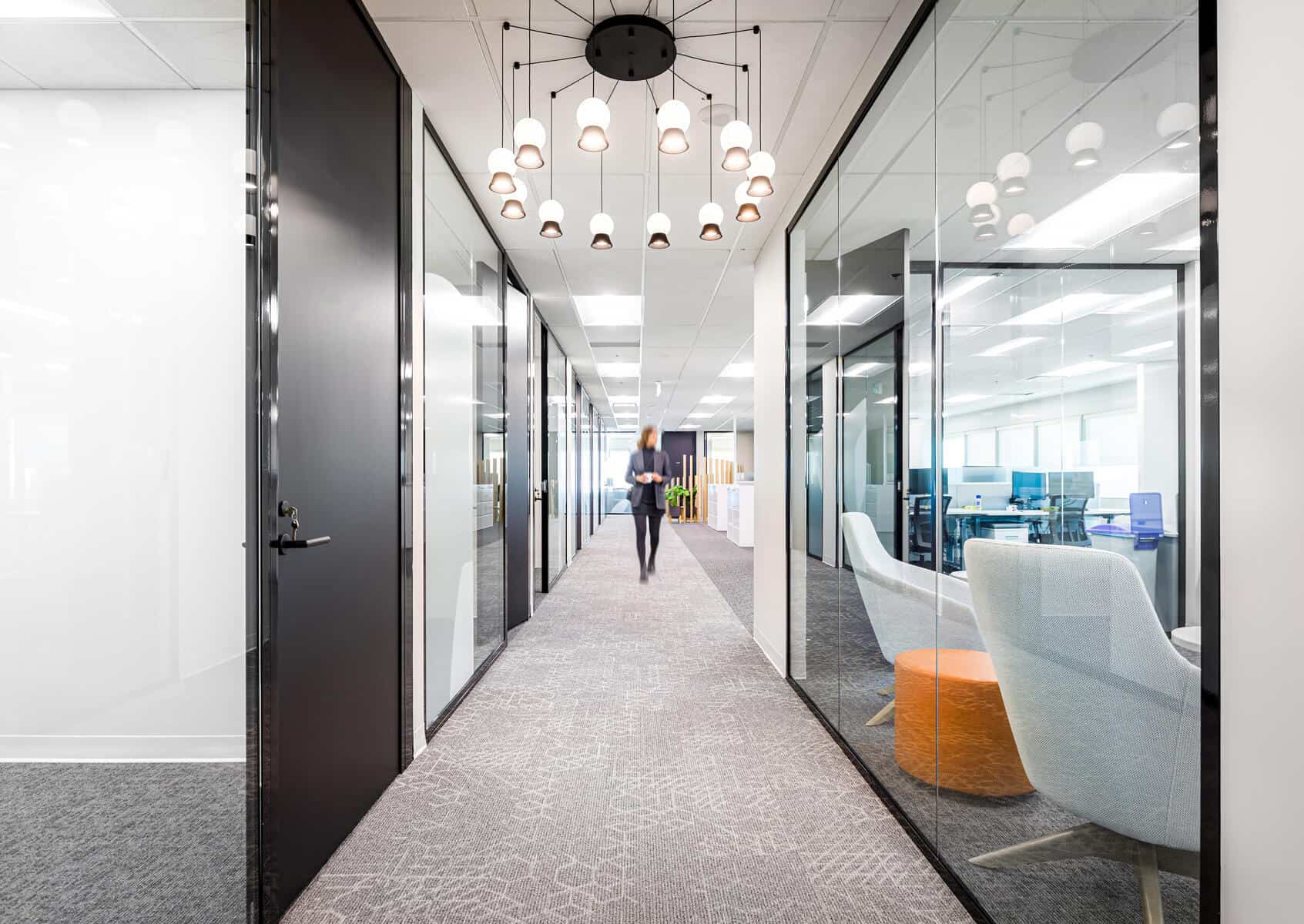 Modern office corridor featuring glass walls, pendant lighting, and a person walking in the distance after an office renovation.