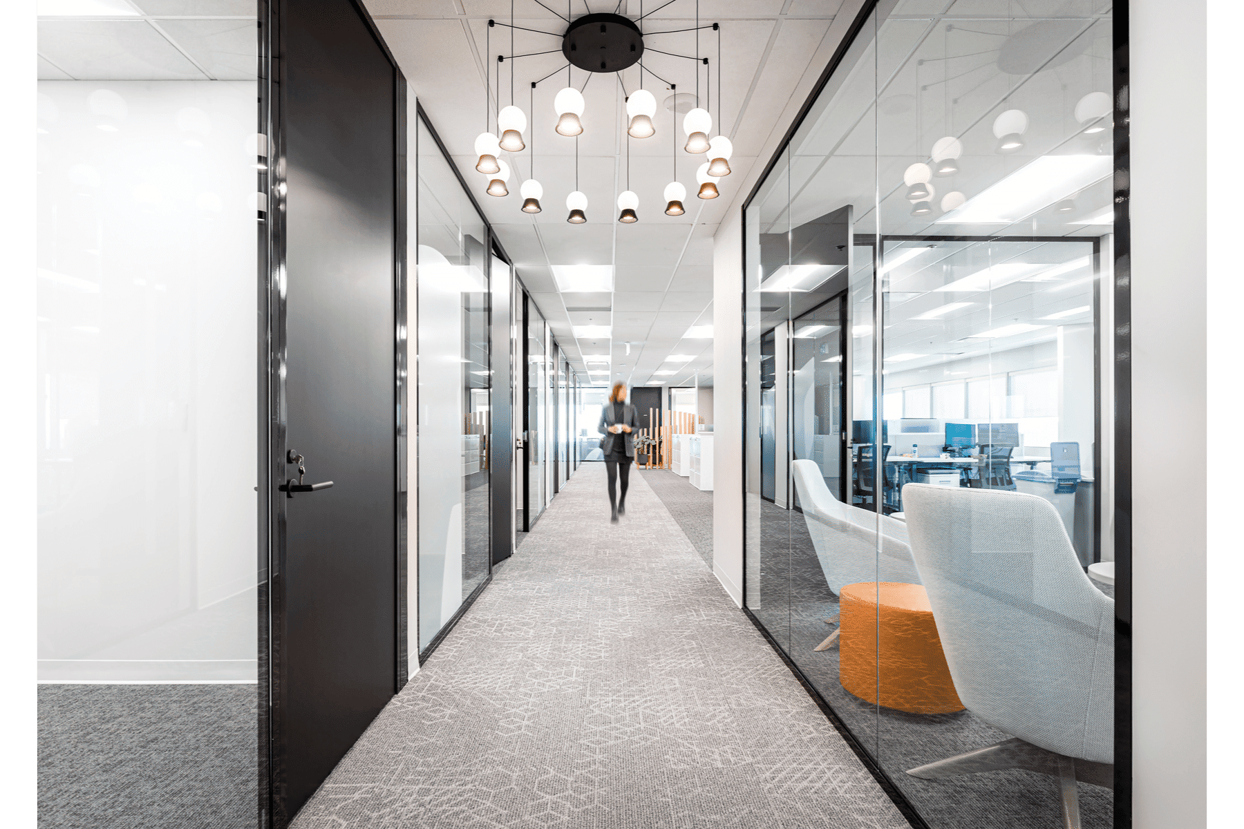 Modern office corridor with glass walls, a person walking in the distance, geometric carpet, and a stylish hanging light fixture.