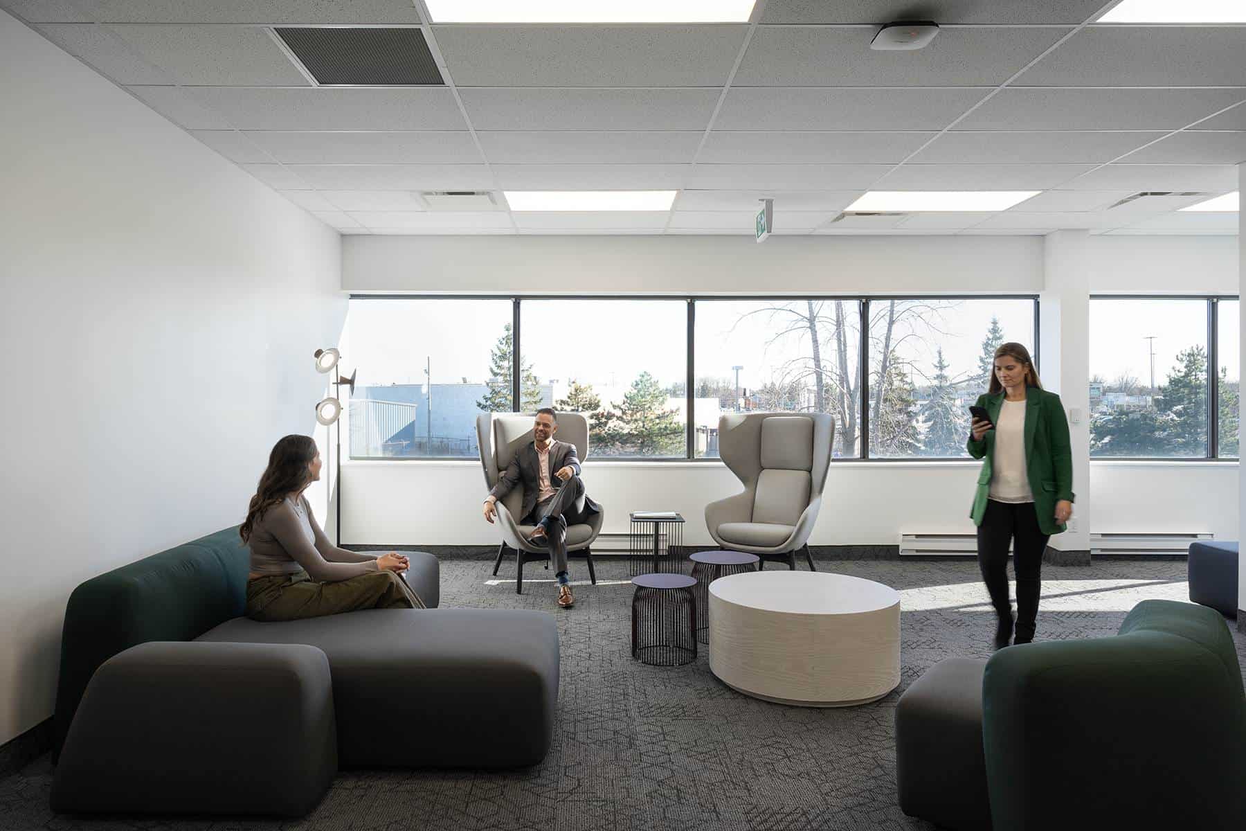 Modern office lounge with three people; two sitting and talking, one walking, near large windows with a view of trees.