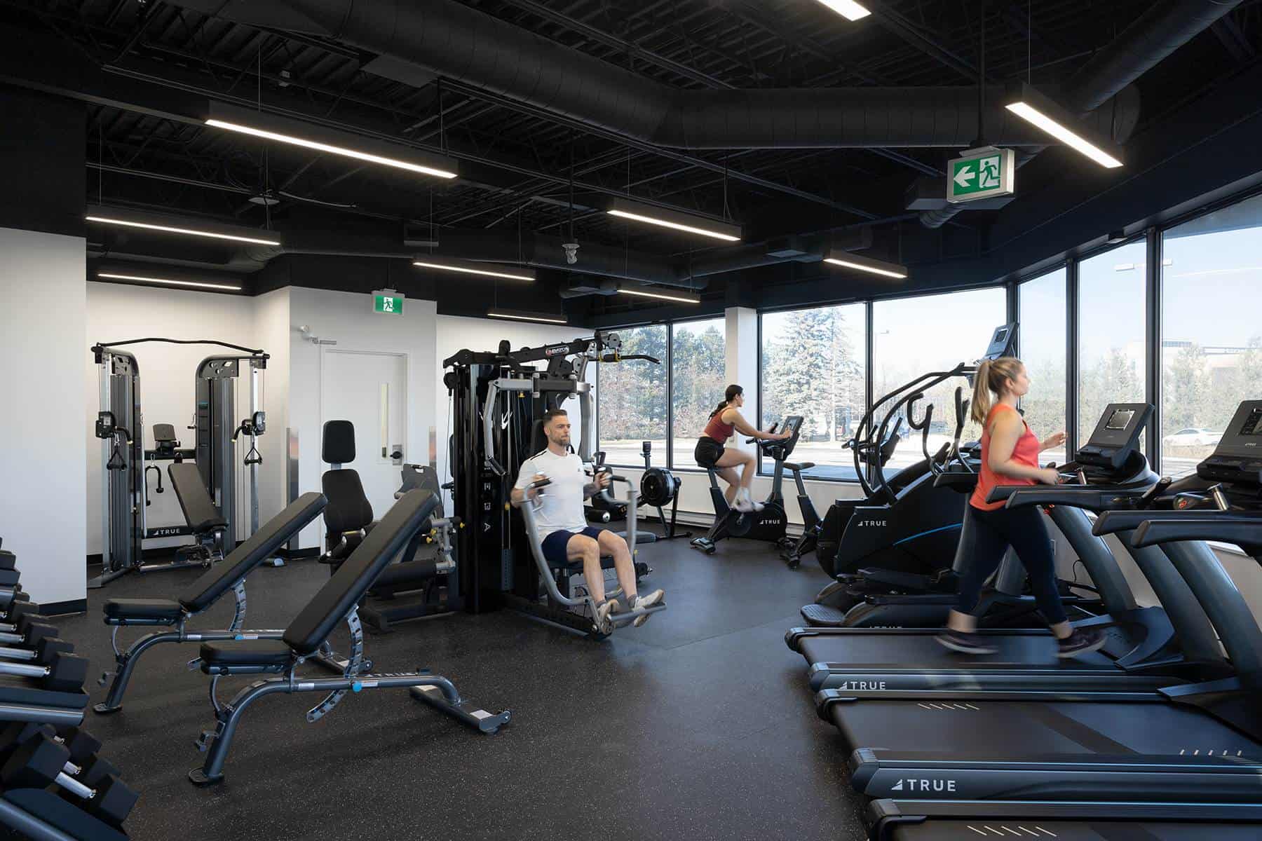 Interior of a modern gym with various exercise machines and people working out, including treadmills and weight stations, redesigned for strategic workplace design to enhance workplace wellbeing.