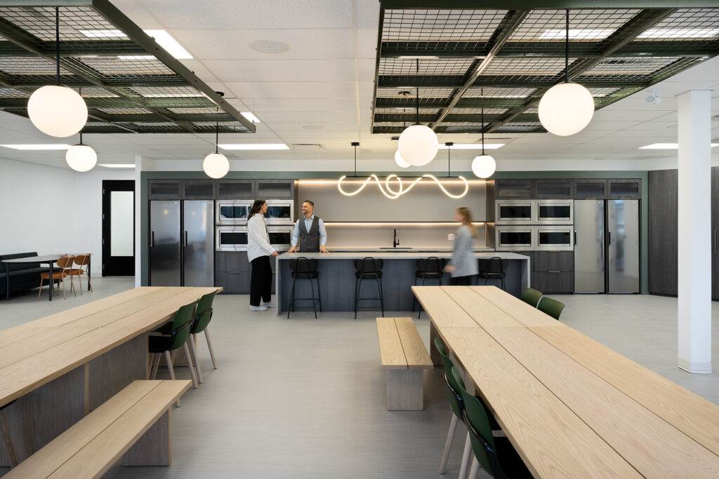 Modern office kitchen and dining area with wooden tables, pendant lights, and employees interacting.