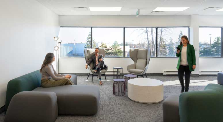 Three people in a modern office lounge, two seated and one standing, engaged in a discussion about a new project in a brightly lit room with large windows.