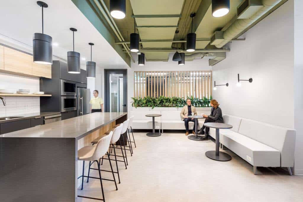 Modern office design and build featuring a kitchen and lounge area with people seated and talking, showcasing stylish interior decor illuminated by natural light, green accents, and communal tables.