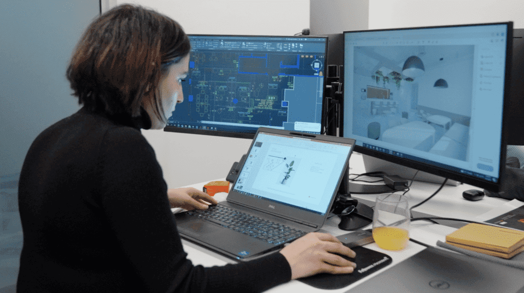 A woman at a desk analyzing architectural designs on multiple computer screens in a design service office setting.