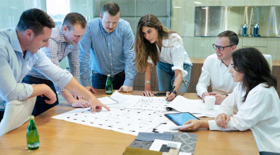 A group of six design service professionals, including three men and three women, collaboratively working over documents on a table in a bright office setting.