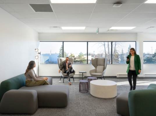 Three professionals in a modern office lounge, two seated and one walking, discussing service design with bright daylight coming through large windows.