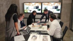 A group of professionals gathered around a conference table, reviewing architectural plans and material samples, with dual screens displaying SEO design presentations in the background.