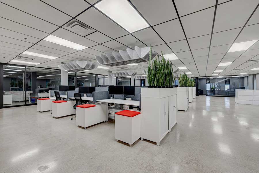 Modern office space with cubicles, red chairs, white desks, and green plants, featuring reflective flooring and overhead lighting as part of a comprehensive workplace design.