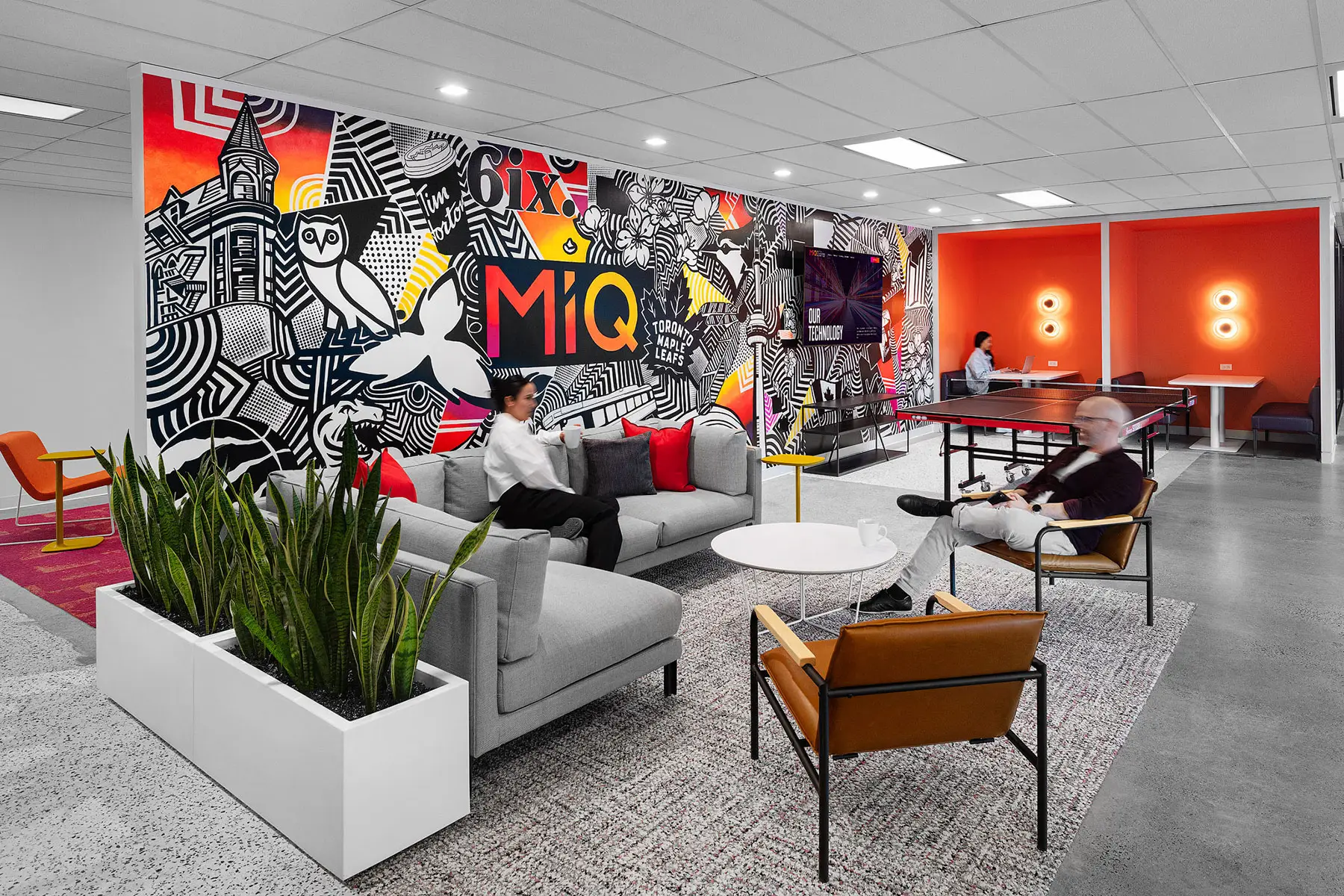 Modern office lounge with colorful graffiti mural, gray sofas, people sitting and chatting, a ping-pong table in the back, and contemporary decor.