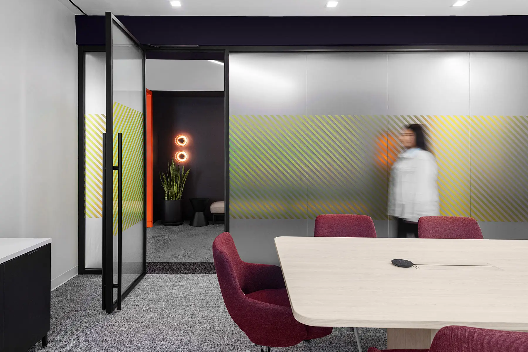 A person in motion walks by a glass partition in a modern office space. The office features a meeting room with a light wood table, red chairs, and decorative lighting.