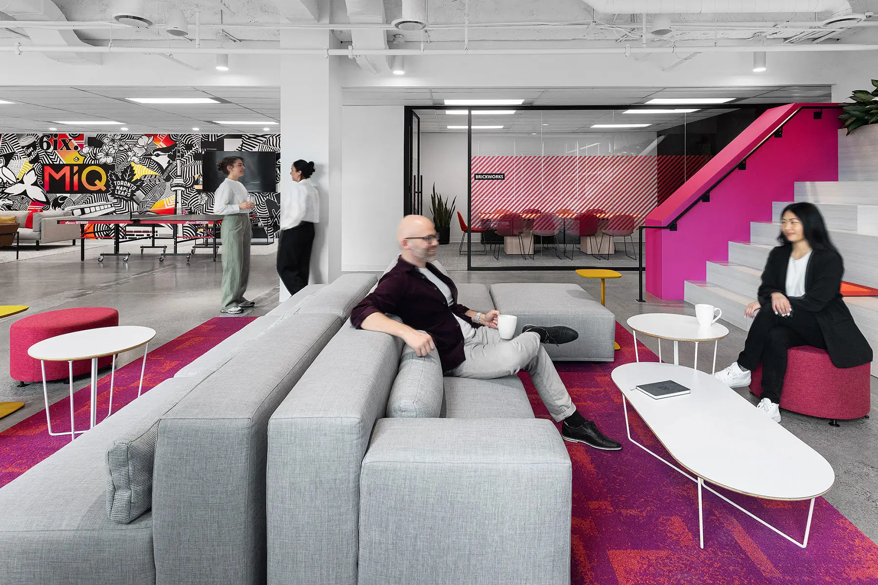 Modern office lounge with grey seating, pink accents, and three people sitting and talking. Two people standing in the background near a brightly decorated wall with "MiQ" text.