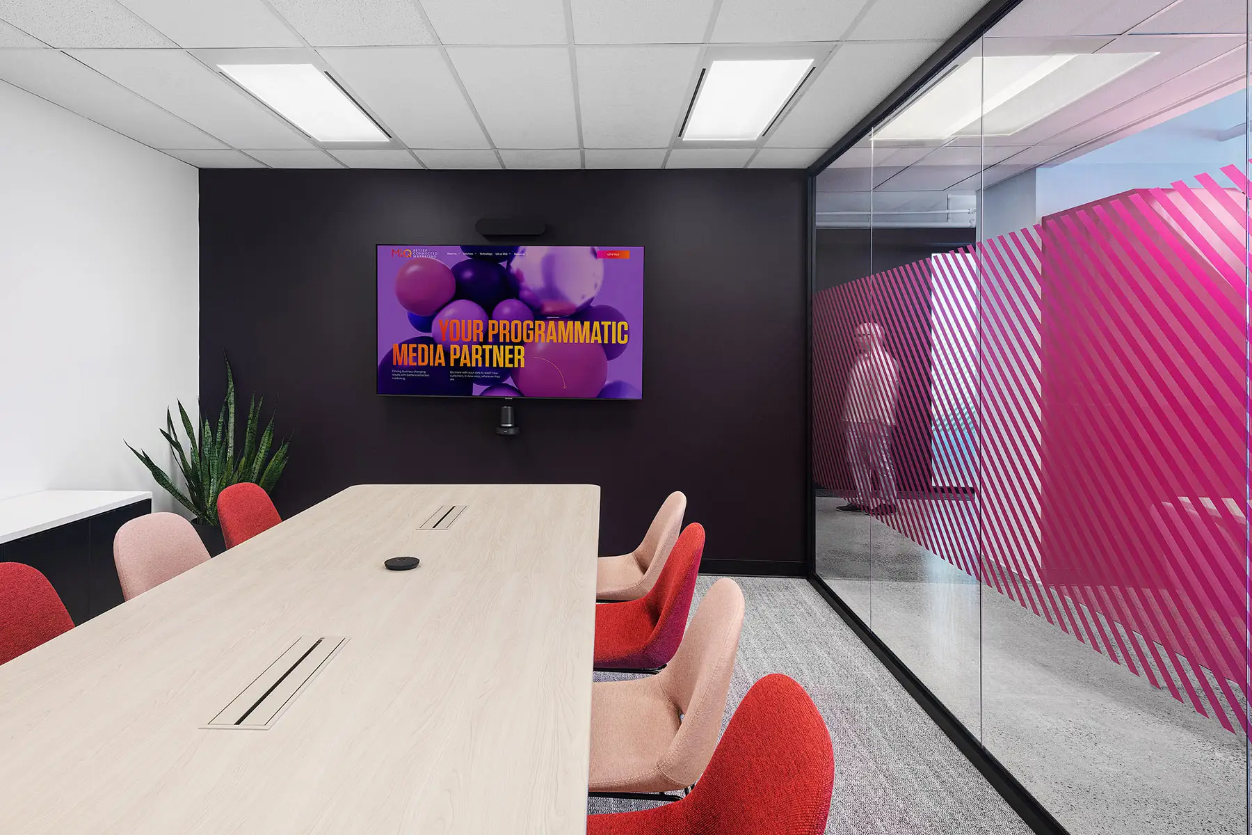 A modern conference room with a long wooden table, red chairs, a large screen displaying "Your Programmatic Media Partner," and glass walls with diagonal pink stripes.