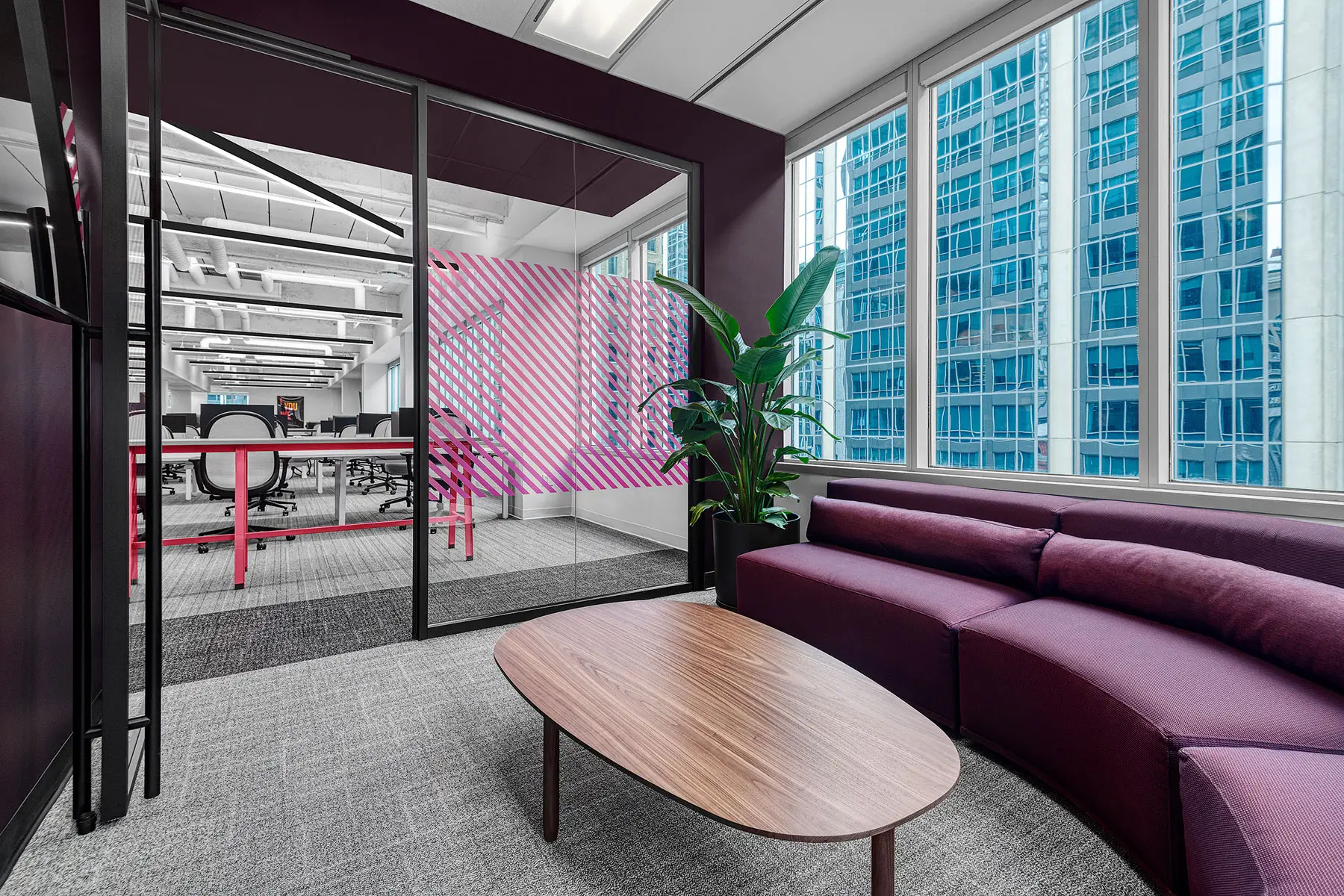 A modern office lounge area with a purple couch, wooden coffee table, and plant, adjacent to a conference room divided by glass walls with pink accents. Tall windows offer a view of neighboring buildings.