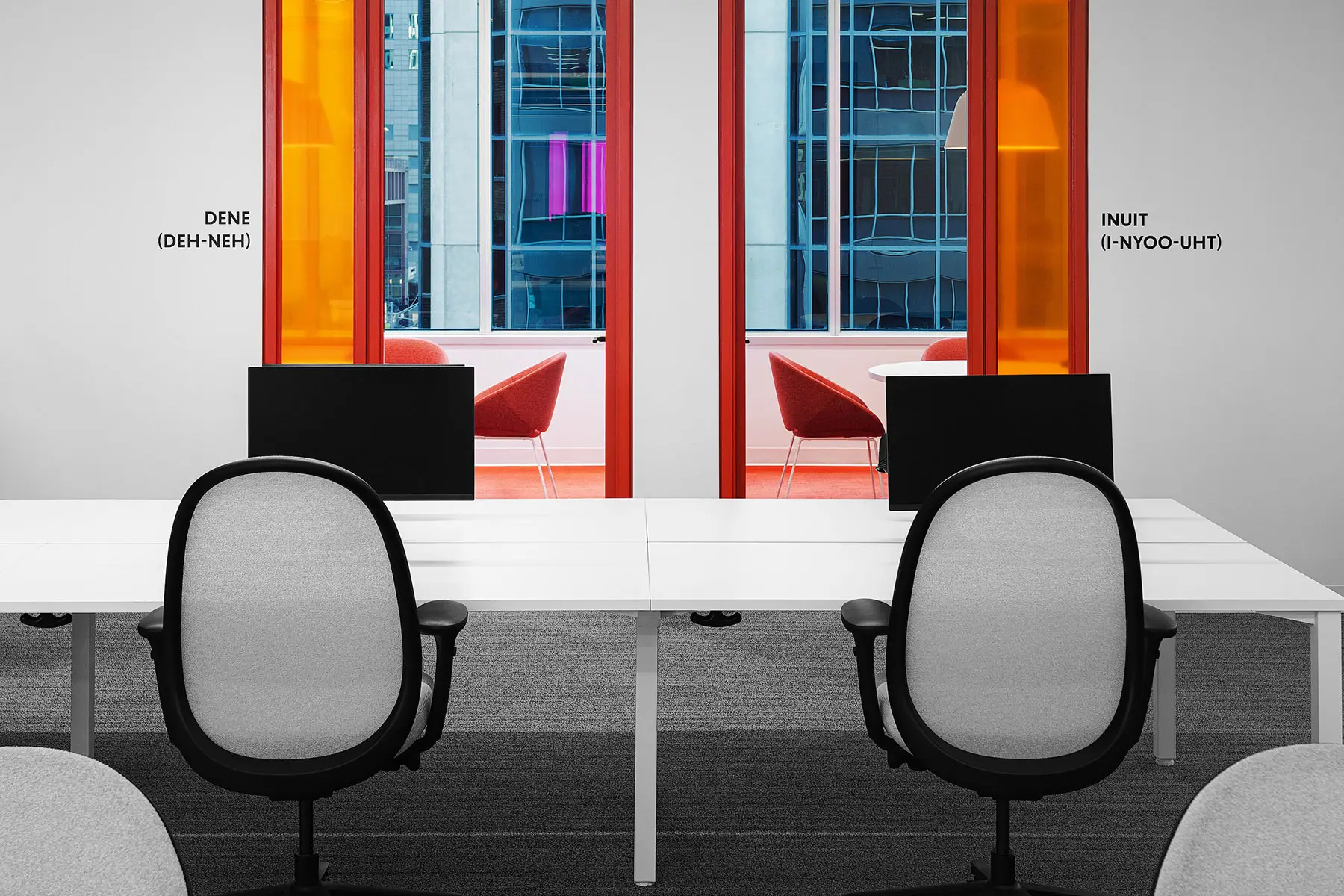 Office space with two desks and two chairs facing two large windows with bright orange frames. Words "Dene (Deh-Neh)" and "Inuit (In-Yoo-Uht)" are labeled above the windows.