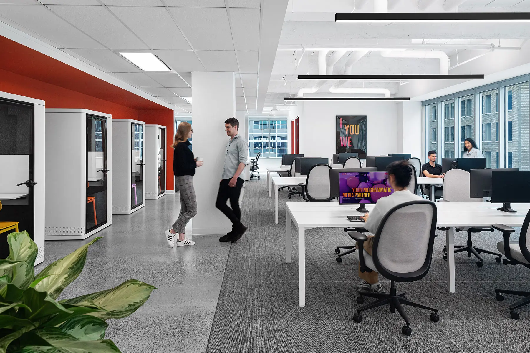 A modern office space with people working at desks and interacting. There are partitioned workstations, large windows, and overhead lighting. A plant is in the corner. One monitor screen is visible.