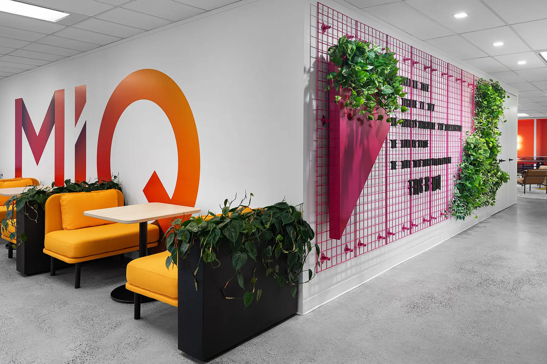 Modern office space featuring yellow seating booths, green plants, a colorful wall art installation, and an open area with tables in the background.