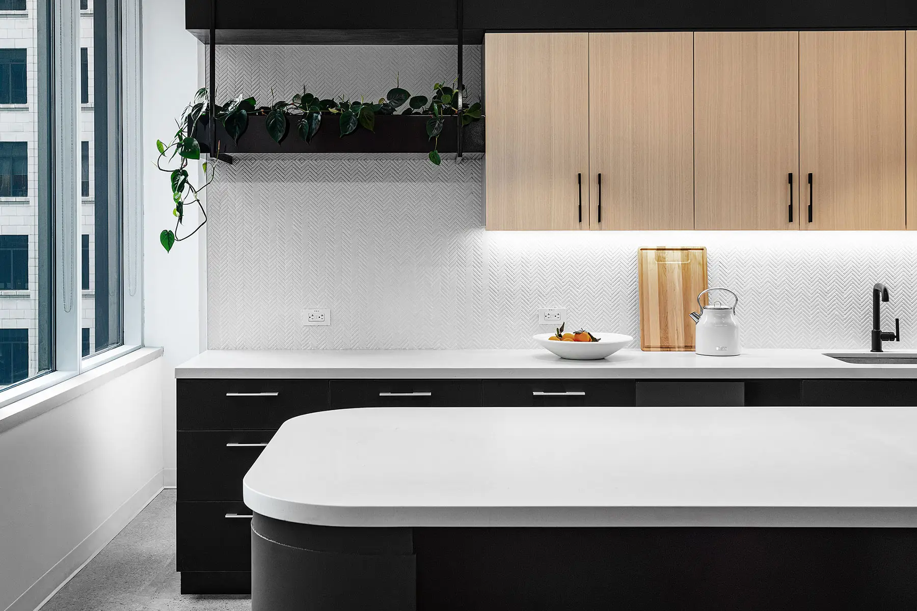 Modern kitchen with a white countertop, black and wood cabinets, and a sink. A bowl of fruit, a kettle, and a wooden utensil holder are on the counter. Potted plants are on a shelf above the cabinets.