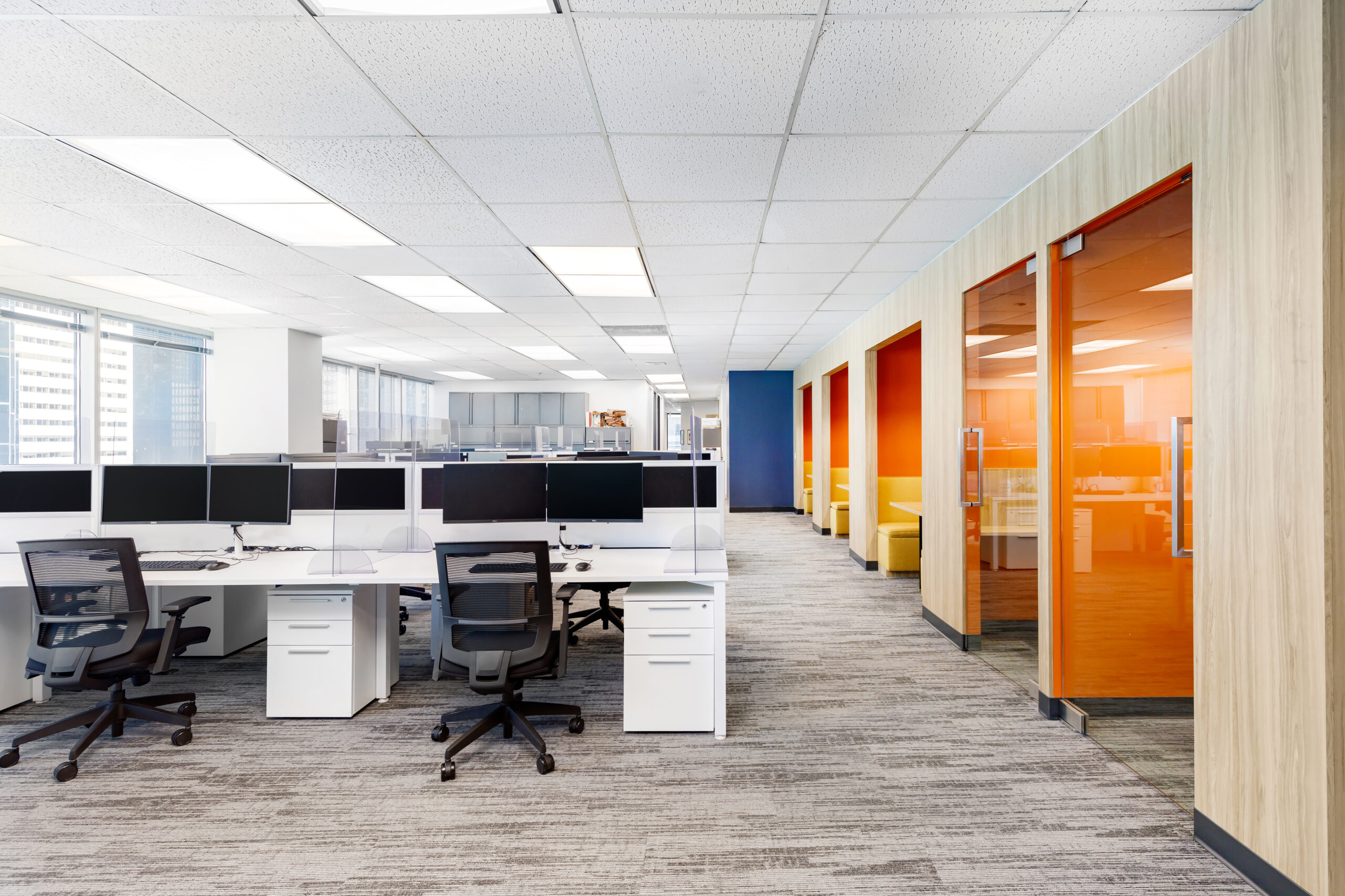 Modern open-plan office with several desks and computer monitors on the left, and glass-walled meeting rooms with orange accents on the right. The space is brightly lit with natural light.