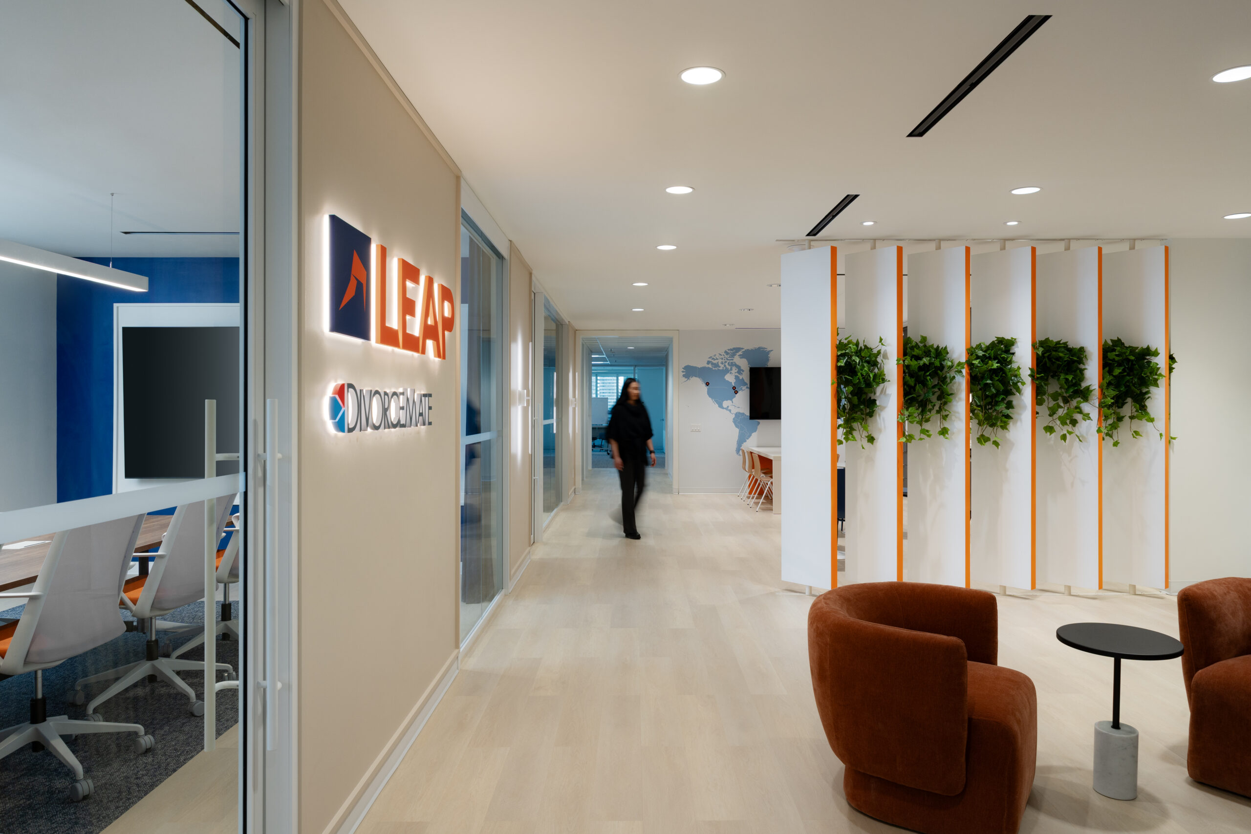 A modern office hallway with a LEAP logo on the wall, a person walking, glass-walled conference room, and seating area with brown chairs and vertical planters.