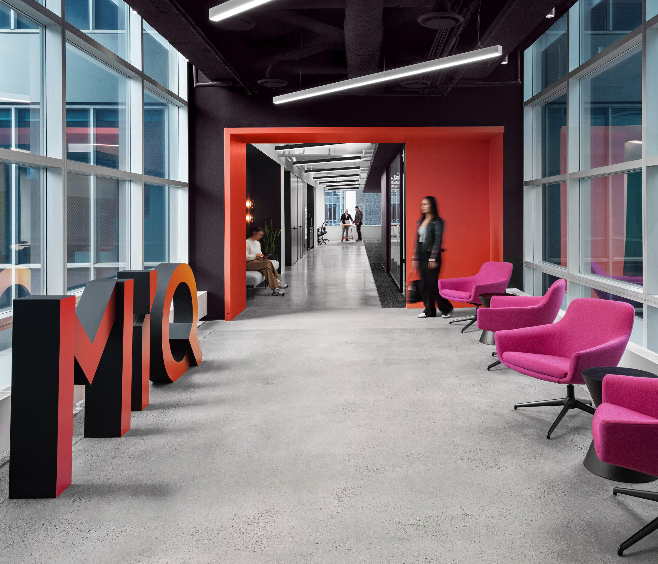 Modern office hallway with large windows, bright pink chairs, and the letters "MiQ" in the foreground. A person walks through the corridor, and others are visible in the background.