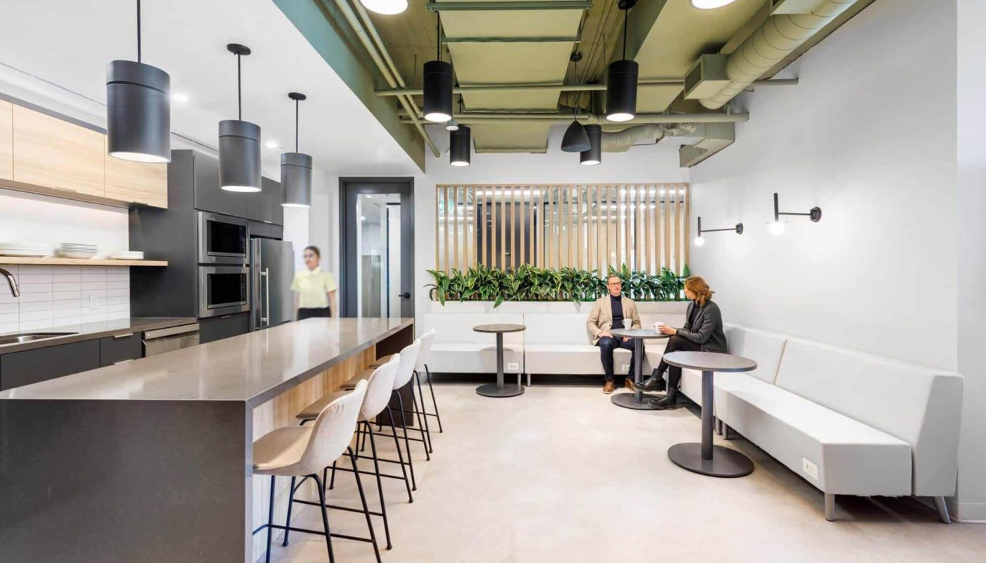 Modern office design and build featuring a kitchen and lounge area with people seated and talking, showcasing stylish interior decor illuminated by natural light, green accents, and communal tables.