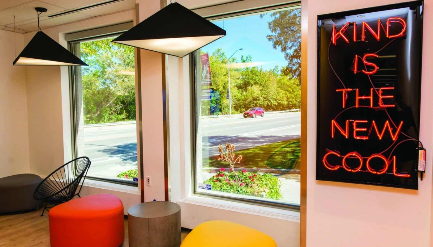 A bright room with large windows, featuring a neon sign that reads "Kind is the new cool." There are colorful ottomans and stylish hanging lamps.