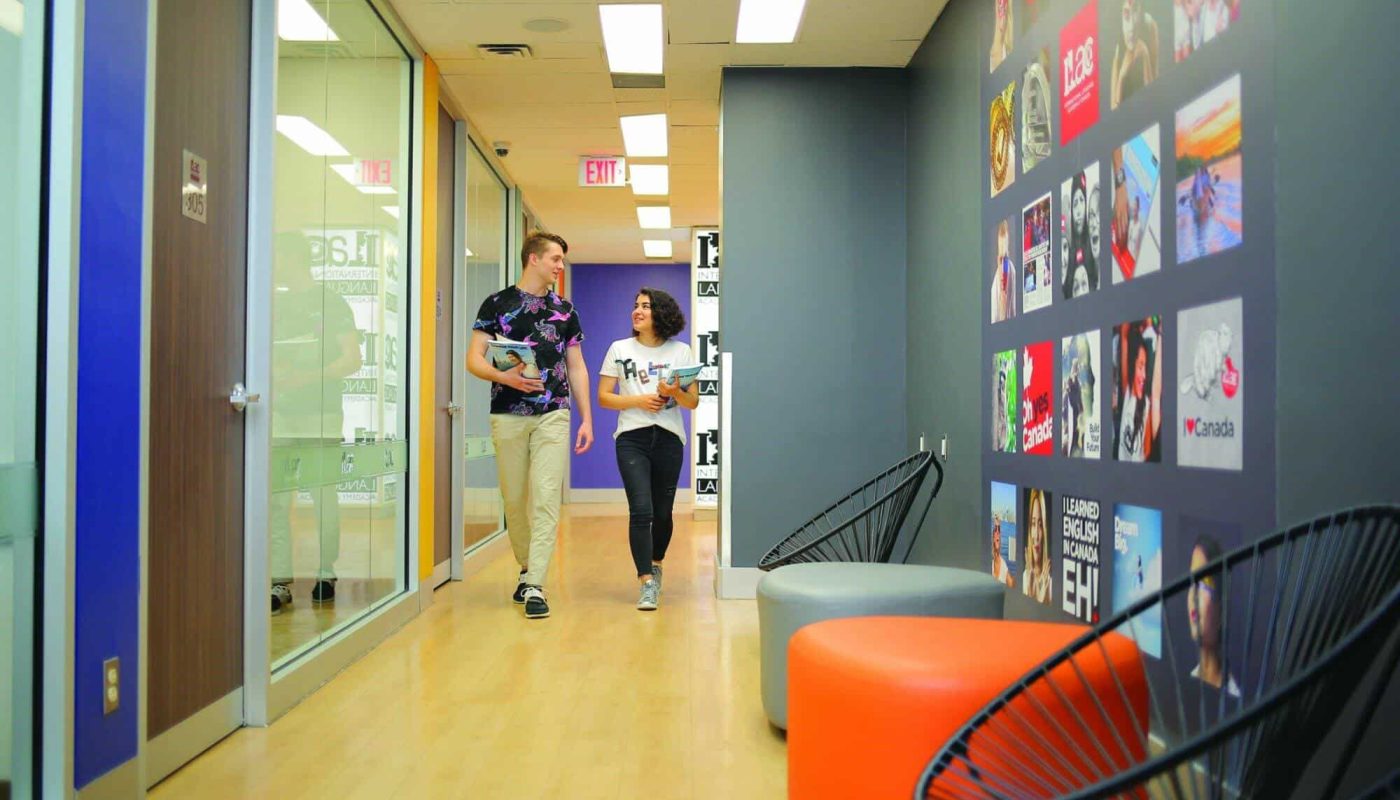 Two people walking and chatting in a brightly lit office corridor with colorful ILAC posters on the wall.