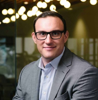 Professional man in glasses wearing a gray suit and blue checkered shirt, smiling confidently on the new Teams Page.