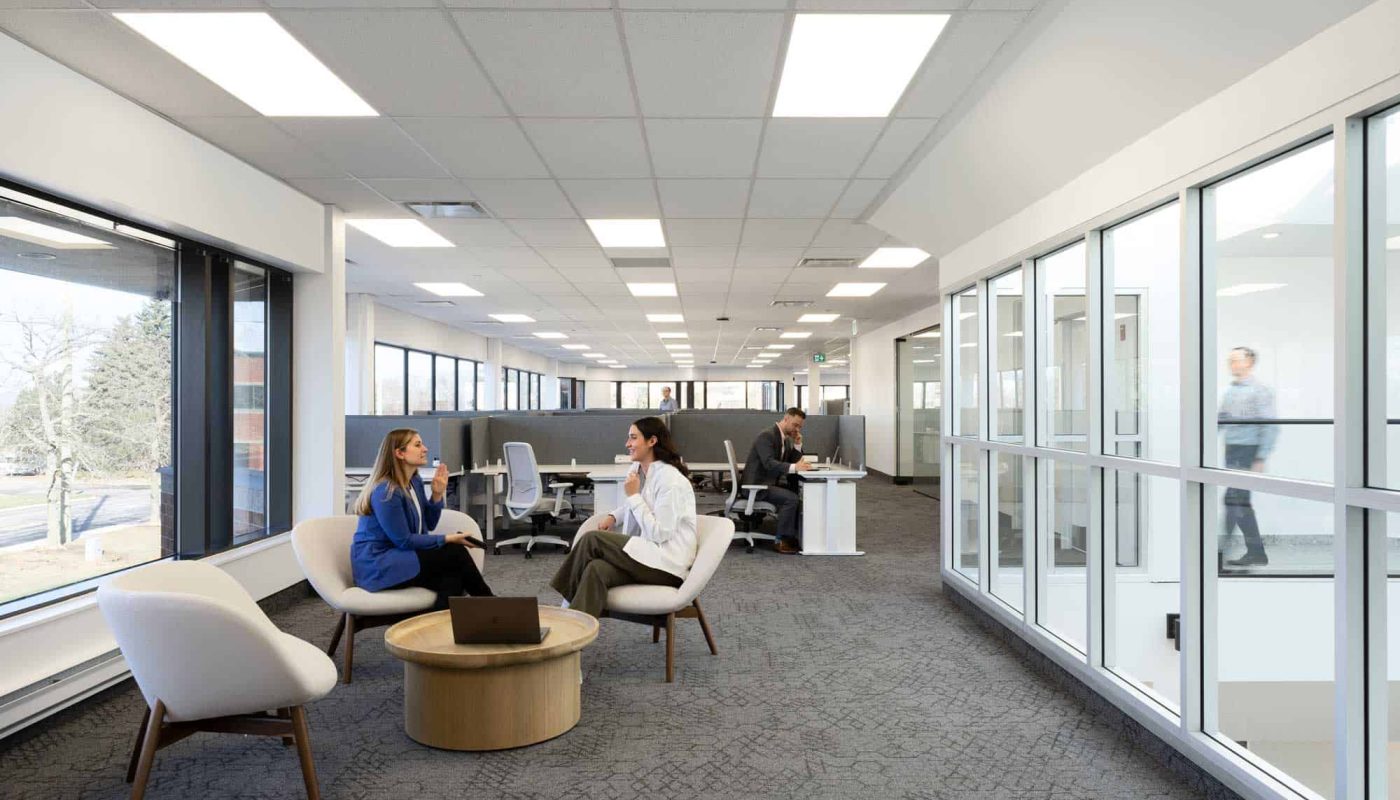 Modern office space with employees at desks and in conversation areas, large windows, and a bright, neutral color scheme featuring commercial office furniture.