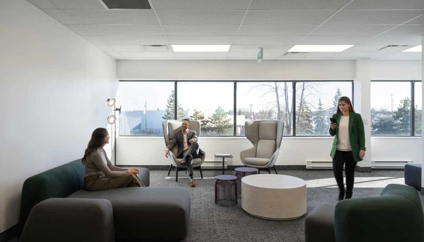 Three people in a modern office lounge with commercial office furniture, one walking and two seated, conversing in a relaxed setting with large windows and neutral decor.